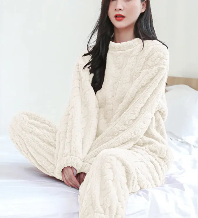 Woman wearing a white -colored cable knit sweater and pants sitting on a bed.